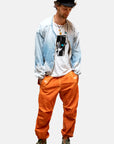 A man in a light blue jacket, white graphic tee, Free City Out of Sight Poplin Jump Pant in orange, green sneakers, layered necklaces, and black cap stands with hands in pockets, looking down.