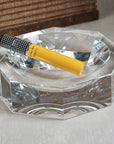 A single yellow cigar in a clear glass tube rests on the Zodax Octagon Bowl, a faceted crystal-like glass ashtray set on a beige textured surface, with brown woven material in the background for a modern look.