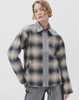 A person with short, wavy blonde hair models the Rag & Bone carrie reversible jacket in muted beige, blue, and gray plaid tones, paired with gray pants, against a plain white background.