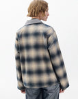 A person with short, wavy blond hair stands with their back to the camera, wearing the Rag & Bone carrie reversible jacket in beige and navy plaid with a gray collar and gray pants against a plain light background.