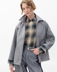 A person with short, wavy blonde hair wears the Rag & Bone carrie reversible jacket over a plaid shirt and high-waisted gray trousers, posing with one hand in their pocket against a plain white background.