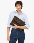 A person with short brown hair, dressed in a light blue shirt and blue jeans, holds the Clare Vivier Carryall Clutch w/ Tabs—a large brown woven bag with a magnetic closure—against a plain white background.