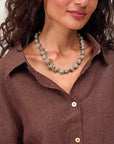 A woman with wavy hair wears a brown pinstriped button-up shirt and the Clare Vivier Checker Shell Necklace. Only her head and upper torso are visible.