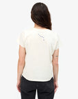 A person with long brown hair faces away, wearing the Clare Vivier Classic Tee CIAO—a lightweight white cotton tee with small blue "CIAO" on the upper back—and black jeans. Their right arm features a small tattoo against a plain white background.