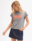 A smiling woman with blonde hair wears the Clare Vivier Classic Tee CIAO, a 100% cotton gray T-shirt with bold orange "CIAO" lettering and short sleeves, paired with a denim skirt, against a plain white background.