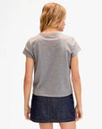 A woman faces away, wearing the Clare Vivier Classic Tee CIAO in gray (100% cotton) with a dark denim skirt. Her blonde hair is loose, she wears a gold bracelet on her right wrist, and the background is plain and light-colored.