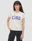 A person with long brown hair wears the Clare Vivier Classic Tee CIAO, a white short-sleeve lightweight cotton T-shirt with bold blue "CIAO" lettering, paired with black pants and standing against a plain light gray background.