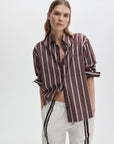 A woman with straight, light brown hair models the Rag & Bone Connie striped shirt, a relaxed-fit cotton poplin top, paired with white pants featuring dark stripes, standing with one hand in her pocket against a plain white background.