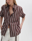 A person wears the Rag & Bone connie striped shirt, a relaxed fit cotton poplin style with white vertical stripes, and off-white pants with a brown side stripe. The shirt is partially unbuttoned with rolled-up sleeves, set against a plain white background.