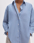 A person wears the Rag & Bone connie striped shirt, a relaxed fit blue and white cotton poplin with vertical stripes, top buttons undone and hands in pants pockets. The image is cropped at the shoulders and thighs.