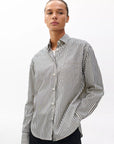 Model wearing the Rag & Bone connie striped shirt, a relaxed fit cotton poplin piece with black and white vertical stripes, stands against a plain white background with one hand in their pocket.