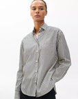 Model wearing the Rag & Bone connie striped shirt, a relaxed fit cotton poplin piece with black and white vertical stripes, stands against a plain white background with one hand in their pocket.