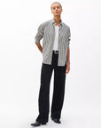 A person stands against a plain white background wearing the Rag & Bone connie striped shirt, a relaxed-fit, black-and-white cotton poplin piece layered over a white top, with black wide-leg pants and shoes. Their hands rest behind their back.