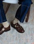 A person sits on a chair, wearing dark blue jeans and Unified Glitter Socks, paired with brown multi-buckle leather shoes, resting their feet on a patterned rug atop a wooden floor.