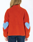 A person wearing the fuzzy orange Coze Magoze jacket by Kerri Rosenthal, featuring large light blue heart elbow patches, stands facing away from the camera against a plain white background.
