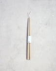 A pair of 18" Taper Candles from The Floral Society, bound together by a white label with minimalist text, exudes a classic design. Set against a light, textured background, these beige candles are attached at the wick ends by a white string loop, promising to bring a graceful ambiance to any setting.