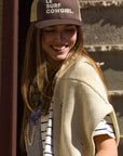A woman in the Ping Pong Surf Club's Le Surf Cowgirl Trucker smiles as she looks down. With long hair, a cream knit shawl, and striped shirt, she stands outdoors in sunlight against a textured wall.