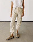 Standing on a concrete floor against a white wall, the person wears a white t-shirt, The Sweatpants by Hey Gang in beige French Terry with an elastic waistband, and light brown clogs.