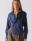 A woman with long blond hair wears The Daily Shirt by Cissa, featuring blue with yellow and white stripes, paired with brown plaid pants against a plain light background.