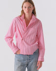 A person with long, light brown hair wears the Cissa Daily Shirt in pink with blue stripes and light blue jeans, posing with one hand in their pocket. Made from soft PAPER COTTON for all-day comfort, set against a plain light background.