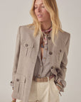 A woman with long blonde hair wears the Maison Hotel Nehru Jacket over a checked shirt, cream eyelet pants, and a patterned scarf, standing with one hand in her pocket and gazing slightly downward and to the side.