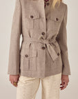 A woman wears the Maison Hotel Nehru Jacket in light beige, belted and with buttoned pockets, over cream eyelet pants. Styled with loose blonde hair and relaxed hands, she stands against a neutral background.