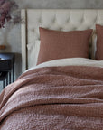A neatly made bed with a tufted headboard features the Pebbled Org Quilt Redwood K by Coyuchi Inc, brown kantha quilts, and two matching pillows. Beside it is a dark nightstand with books, a clock, and dried flowers in a vase. Soft light filters through a window in the background.