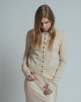 A woman with long blonde hair wears the Brazeau Tricot Royal All Thumbs Cardigan in beige, featuring a button front, paired with a light beige skirt. She stands hands in pockets against a plain light background, looking down.