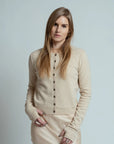 A woman with long blonde hair and a neutral expression stands against a plain light background, wearing the Brazeau Tricot Royal All Thumbs Cardigan in beige with a button front, paired with a matching skirt.