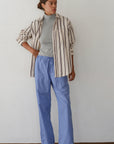 A person with short hair stands casually against a plain wall wearing Donni’s The Stripe Pop Drawstring Pant, a loose striped Cotton Poplin button-up shirt over a gray turtleneck, and dark shoes.