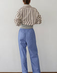 A person stands facing away from the camera, wearing Donni's The Stripe Pop Drawstring Pant in light blue with a cropped fit striped beige shirt and dark shoes, against a plain white wall and brown floor.