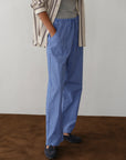 Wearing Donni’s The Stripe Pop Drawstring Pant in blue and white stripes with a cropped fit, a grey shirt, beige and brown striped overshirt, and dark loafers, the person stands on a brown floor against a light wall. Their face is unseen.