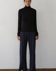 A person stands against a plain white wall wearing Donni's The Baby Rib Turtleneck, loose-fitting dark blue ribbed pants, and beige shoes, facing forward with a neutral expression.
