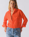 A woman wears The Daily Shirt by Cissa—a bright orange PAPER COTTON eyelet crop with rolled-up sleeves—paired with light blue jeans against a plain background, creating an effortless daily style statement.