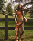 Hunter Bell NYC Allyne Skirt with fringe in rust, styled outdoors with a tank top and sandals by a wooden fence.