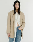 A person with long black hair stands against a plain white background, wearing Denimist's DECONSTRUCTED BLAZER WITH BELT in beige, styled over a white striped shirt and blue jeans.