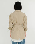 A person with long dark hair, seen from behind, is wearing the Denimist DECONSTRUCTED BLAZER WITH BELT in beige over a white shirt and blue jeans, standing against a plain white background.