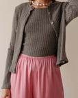 A person wears Donni’s The Sweater Rib Cardi with ribbed texture and mother of pearl buttons, layered over a matching top, styled with loose light pink pants and a chunky pearl necklace. Their face is not visible.