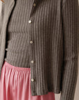 A person wears Donni's The Sweater Rib Cardi in gray with mother of pearl buttons over a matching ribbed top, paired with pink satin pajama pants. Only the torso and neck show, highlighting the cardi’s cozy ribbed texture.