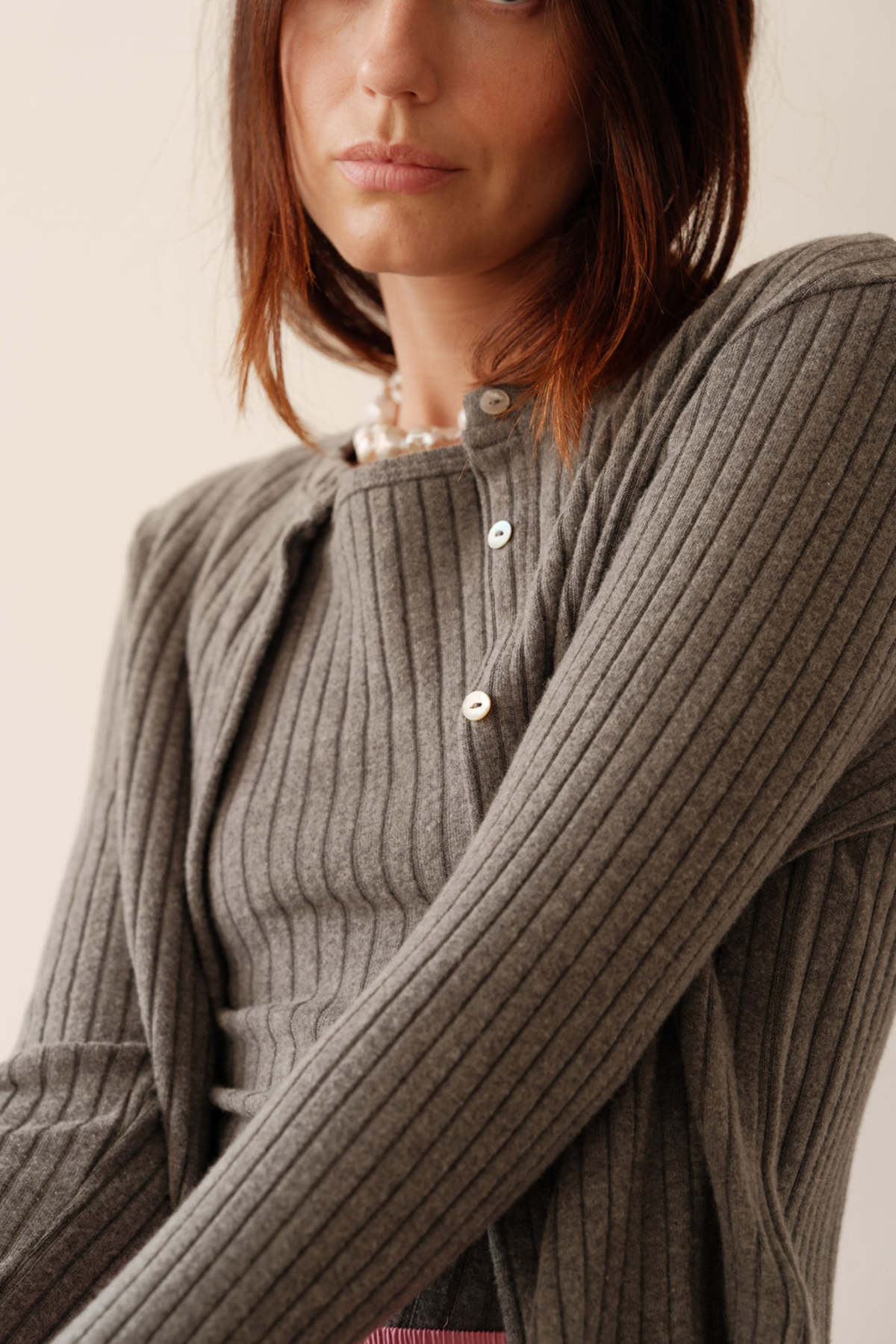 A woman with straight brown hair models Donni's The Sweater Rib Cardi in grey, featuring ribbed texture and mother of pearl buttons. She gazes slightly away from the camera, pictured chest up against a plain light background.