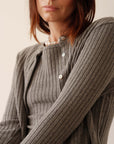 A woman with straight brown hair models Donni's The Sweater Rib Cardi in grey, featuring ribbed texture and mother of pearl buttons. She gazes slightly away from the camera, pictured chest up against a plain light background.