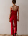 A person with a butterfly tattoo on their upper back stands facing away, wearing Donni's red The Rib Kick Flare tank top and matching pants, showcasing the high-rise fit. They are on a textured rug against a plain wall backdrop.