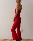 In a minimalist room, a person barefoot and slightly turning glances over their shoulder, wearing the Donni Rib Kick Flare pants with a high-rise fit and a bright red sleeveless top. The floor features a woven texture.