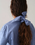 A person with long, wavy brown hair is seen from behind, wearing a light blue shirt and securing their ponytail with Donni's The Taffeta Bow Cornflower Blue accessory.