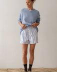 A person stands indoors against a plain background, wearing Donni's light blue sweater paired with The Pop Boxer shorts, crafted in Los Angeles. Their hair is tied back and they wear socks with brown shoes, creating a relaxed and effortlessly composed look.
