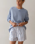 Sporting Donni's The Pop Boxer, a person in a light blue sweater and white cotton poplin shorts with blue stripes poses against a plain backdrop. Made in Los Angeles, the relaxed fit enhances their serene expression as they lightly grasp the waistband of their shorts.