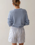 Facing away, a person with a bun hairstyle wears The Pop Boxer by Donni—white shorts with blue vertical stripes—and a light blue cotton poplin sweater. Made in Los Angeles, the ensemble is styled against a neutral background for a simple, minimalist look.