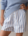 A person wearing Donni's The Pop Boxer in white stripes and a light blue, relaxed fit sweater stands with one hand on their hip. The sweater is slightly tucked into the cotton poplin shorts, which are made in Los Angeles and feature button details.