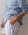A person wearing The Pop Boxer shorts by Donni, featuring blue vertical stripes on white, and a light blue sweater, exudes a relaxed fit with one hand tucked in the pocket. Made from cotton poplin, they stand effortlessly against a plain light background.