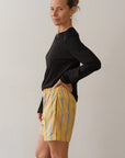 With their hair tied back, a person stands sideways and looks at the camera, wearing Donni's The Pop Boxer shorts in yellow with blue and white stripes, alongside a relaxed fit black long-sleeve shirt. They rest one hand on their hip against a plain, light backdrop.